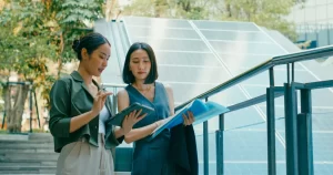 Duas profissionais analisam documentos ao lado de uma estrutura com painéis solares em área externa, sugerindo trabalho com energia renovável e tecnologia limpa.